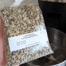 A clear plastic bag filled with whole green coffee beans, featuring a white label with text. In the background, a metal coffee brewing vessel is visible.